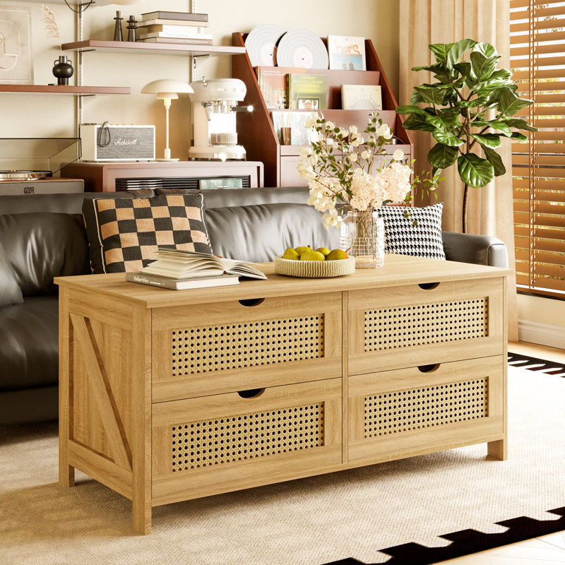 Natural wood coffee table with four storage drawers, featuring rattan panels. Decorated with flowers, a fruit bowl, books, and checkered cushions.
