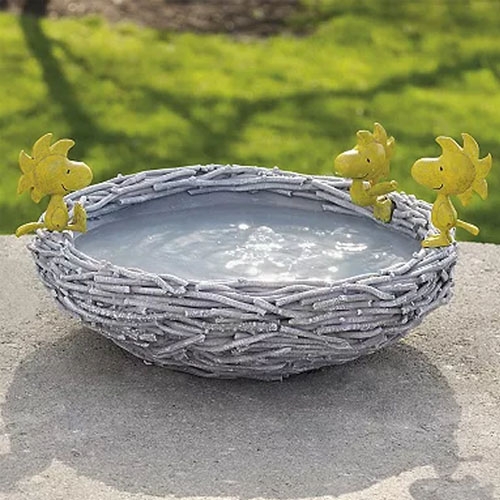 A birdbath designed like a woven nest, featuring three yellow characters resembling Woodstock perched on the rim. The bowl is filled with water, set outdoors on a concrete surface amidst green grass.