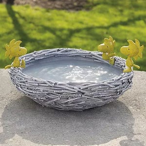 A birdbath designed like a woven nest, featuring three yellow characters resembling Woodstock perched on the rim. The bowl is filled with water, set outdoors on a concrete surface amidst green grass.