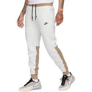 White and beige Nike Tech joggers feature a tapered design with a drawstring waist, side pockets, and a small black Nike logo on the left thigh.