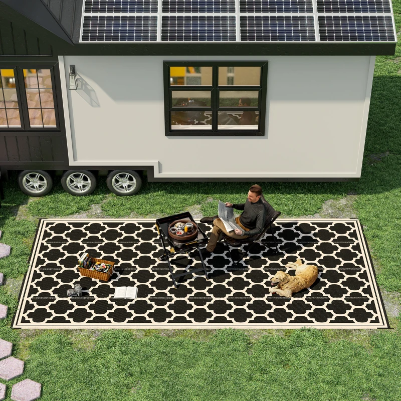 A tiny house on wheels features solar panels on the roof. A person is sitting on a patterned outdoor rug with a dog nearby, a small grill table, and a basket on the grass.