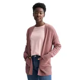 A woman is wearing a light pink t-shirt, a dusty pink open cardigan, and blue jeans. Her hands are in the cardigan pockets, and she stands against a plain white background.