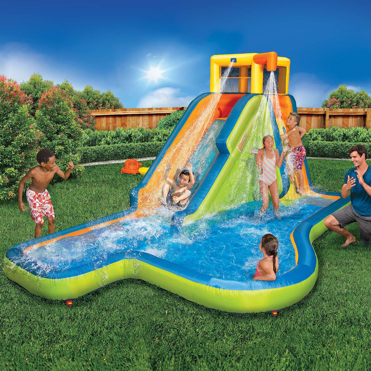 Inflatable water slide with a climbing wall, splash pool, and two water sprinklers in a backyard setting, featuring children and an adult enjoying the play structure.