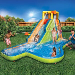 Inflatable water slide with a climbing wall, splash pool, and two water sprinklers in a backyard setting, featuring children and an adult enjoying the play structure.