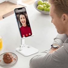 A phone holder allows hands-free video calling, securely holding the phone in any orientation and height. A man sits at a table with juice, a muffin, and a bowl of apples nearby.