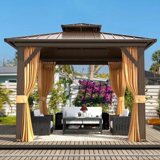 A gazebo with a metal roof and tan curtains is set up on a wooden deck. Inside, there are wicker-style chairs and a coffee table with a decorative item on top.