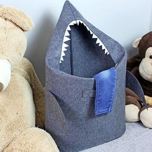 A felt shark-shaped laundry basket with a denim pocket on the side, surrounded by plush toys.