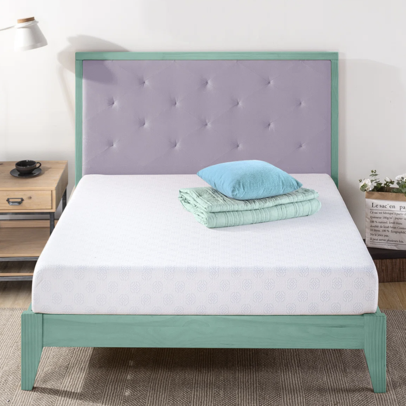 A bed features a tufted headboard and green frame, topped with a folded mint blanket and blue pillow. A bedside table holds a lamp and cup, with a plant in a paper bag nearby.