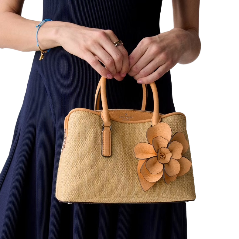 A woven handbag with tan leather trim features a large floral embellishment. A person in a black dress holds the bag, adorned with a blue bracelet.