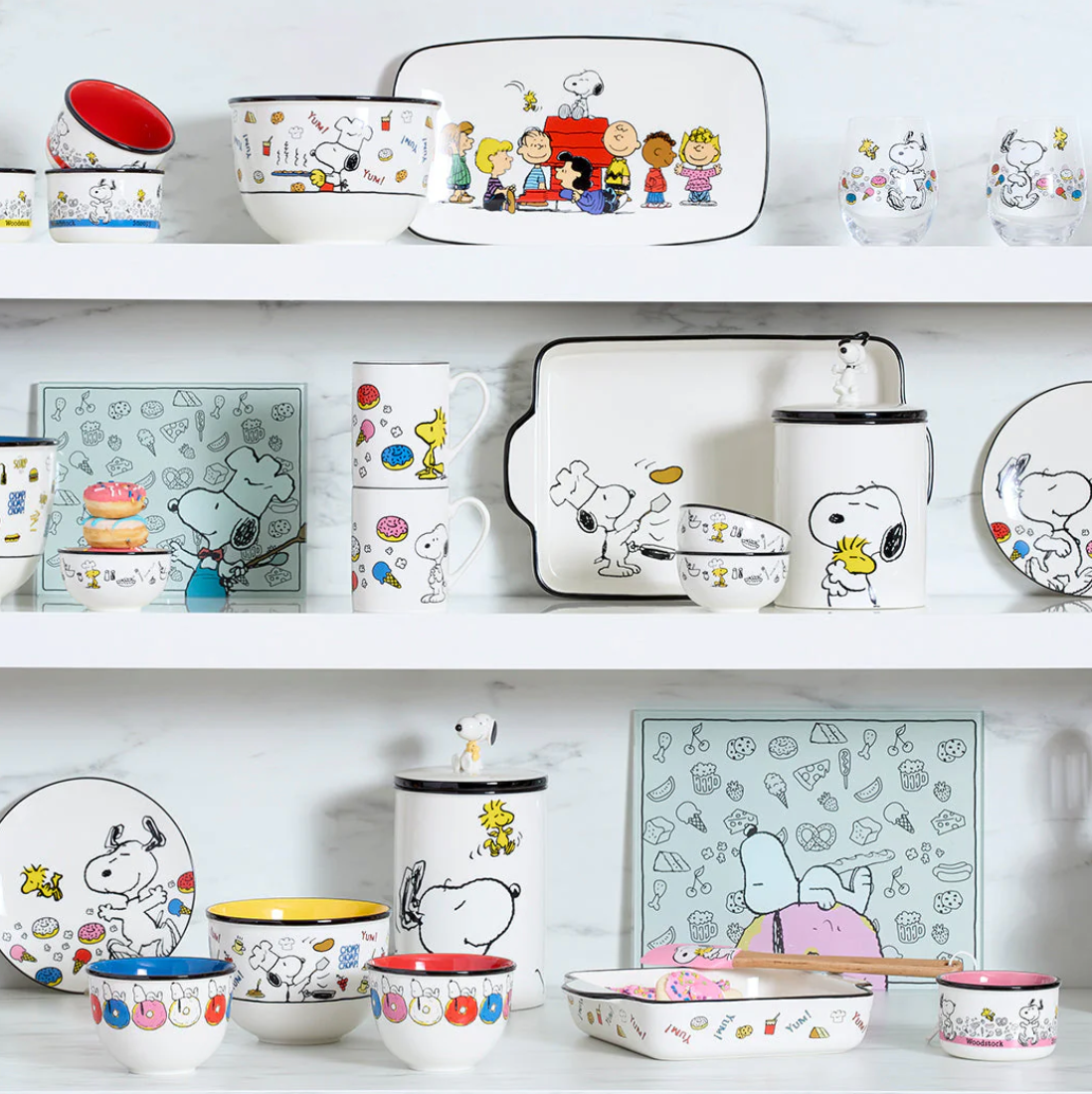 A shelf display featuring Peanuts-themed dishware, including bowls, mugs, plates, glasses, and serving trays with Snoopy and characters in playful designs.