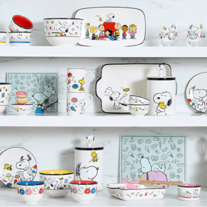 A shelf display featuring Peanuts-themed dishware, including bowls, mugs, plates, glasses, and serving trays with Snoopy and characters in playful designs.