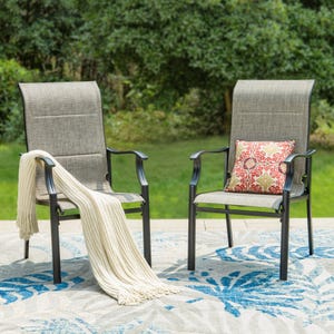 Two patio chairs with padded, grayish fabric seats and black metal frames. One chair has a white knit throw, and the other features a red and white floral cushion, placed on a blue and white patterned outdoor rug.
