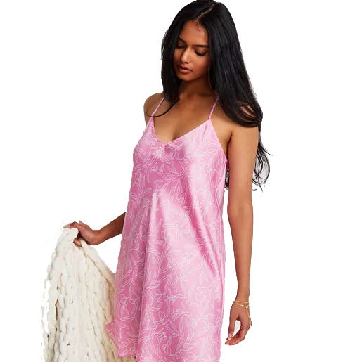 A woman in a pink slip dress with thin straps and a subtle floral pattern holds a chunky knit blanket in one hand.