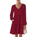 A woman is wearing a long-sleeve, V-neck, dark red tiered dress with elastic cuffs and pockets, accessorized with a delicate necklace and carrying a small woven crossbody bag.