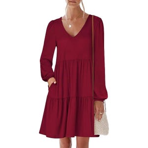 A woman is wearing a long-sleeve, V-neck, dark red tiered dress with elastic cuffs and pockets, accessorized with a delicate necklace and carrying a small woven crossbody bag.