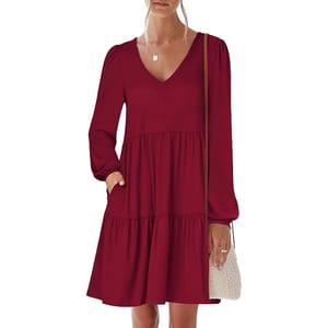 A woman is wearing a long-sleeve, V-neck, dark red tiered dress with elastic cuffs and pockets, accessorized with a delicate necklace and carrying a small woven crossbody bag.