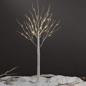 A slender, artificial birch tree with integrated LED lights along its branches, set against a dark backdrop and standing on a simulated snowy surface.