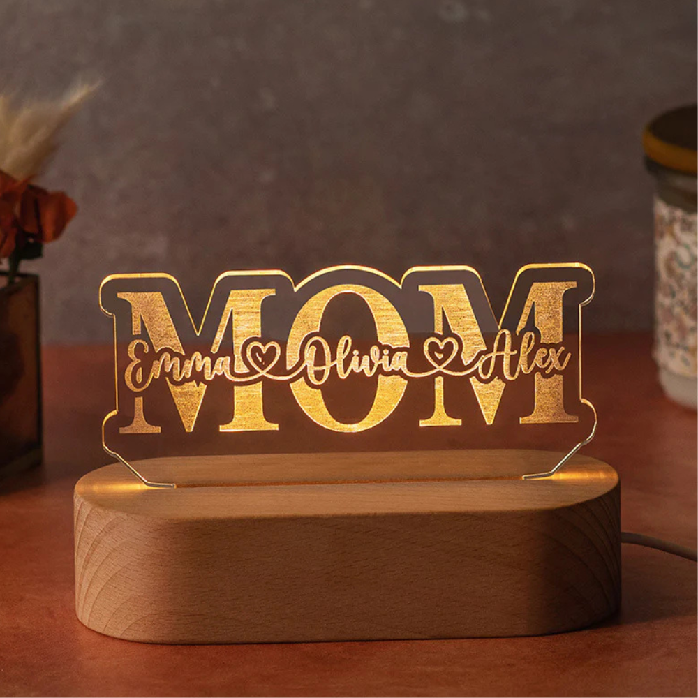 A decorative LED light with the word \“MOM\“ features personalized names \“Emma,\“ \“Olivia,\“ and \“Alex\“ written stylishly across it. It is mounted on a wooden base.
