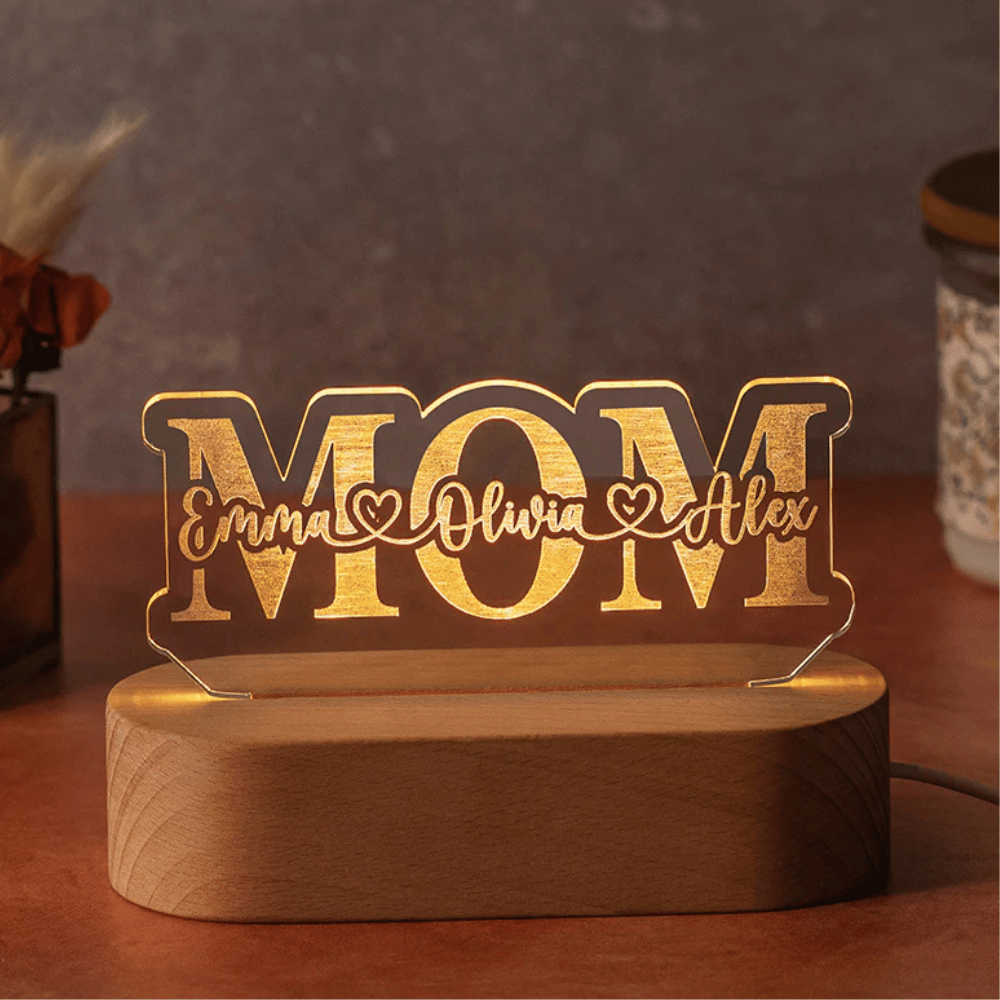 A decorative LED light with the word \“MOM\“ features personalized names \“Emma,\“ \“Olivia,\“ and \“Alex\“ written stylishly across it. It is mounted on a wooden base.