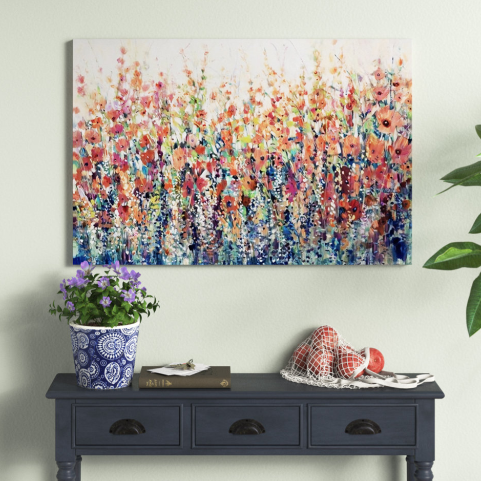 Colorful abstract floral painting on a light wall above a dark blue console table, which holds a potted plant, books, and a net bag with oranges.