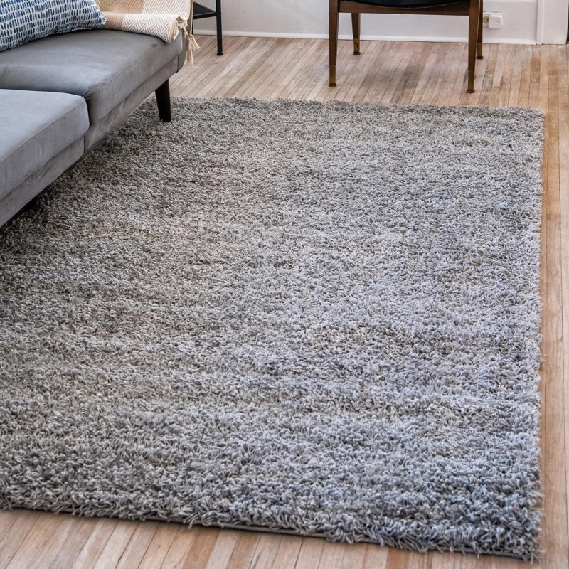 A 5' x 8' gray shag rug with a thick, plush texture is placed on a light wooden floor beside a gray sofa and a wooden chair.