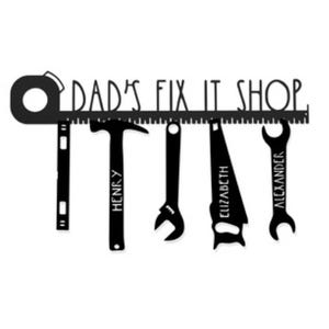 Metal wall art features the text \“Dad's Fix It Shop\“ above a ruler with hanging tools: a level, hammer, wrench, hacksaw, and open-end wrench, each personalized with names \“Henry,\“ \“Elizabeth,\“ and \“Alexander.\“