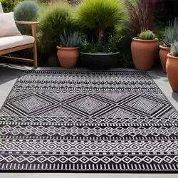 Black and white geometric outdoor rug with diamond patterns placed on a patio, surrounded by potted plants and a wooden bench.