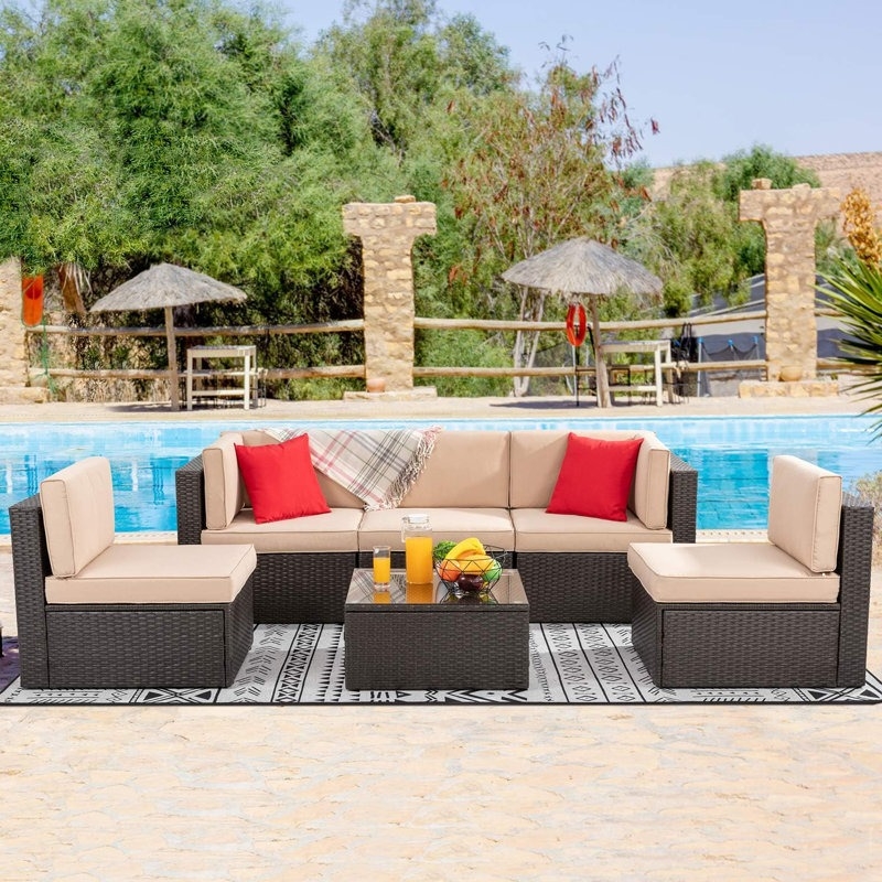 Outdoor patio set with beige cushions and red pillows includes a sectional sofa and a glass-topped coffee table. The setting is poolside with a patterned rug and drinks on the table.