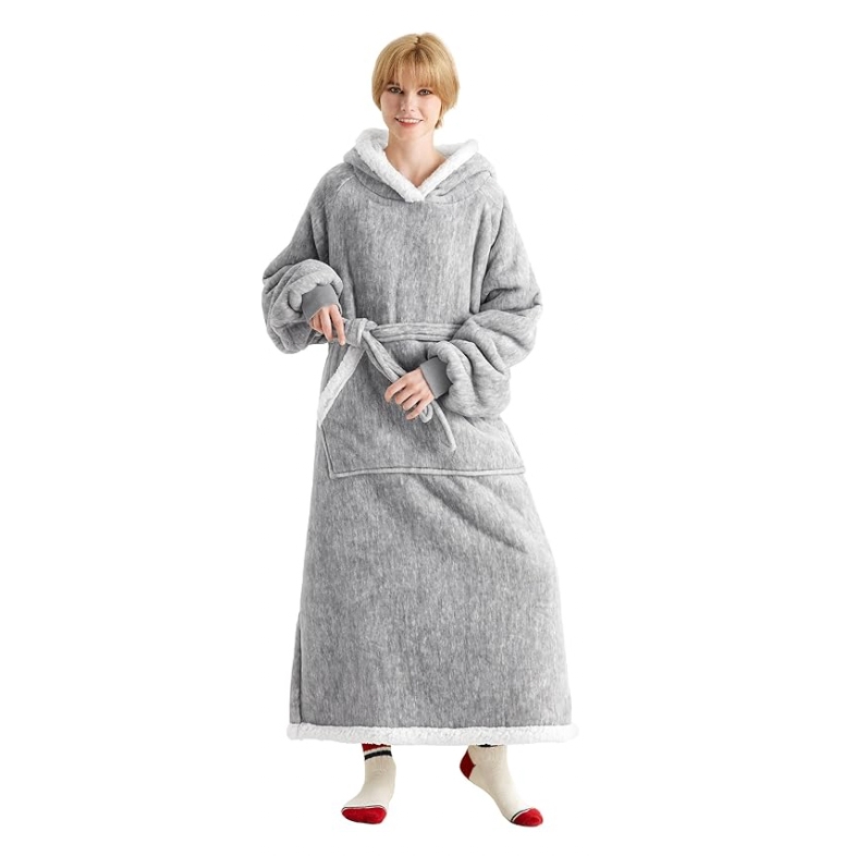 A woman is wearing a gray long blanket hoodie with a front pocket, featuring a hood and sherpa-style lining. The sleeves are oversized with cuffs, and it has a tie belt at the waist. She is wearing red-tipped socks.