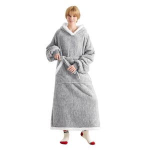 A woman is wearing a gray long blanket hoodie with a front pocket, featuring a hood and sherpa-style lining. The sleeves are oversized with cuffs, and it has a tie belt at the waist. She is wearing red-tipped socks.