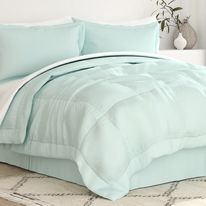 A light blue bedding set includes a comforter, two pillows, and a bed skirt, all displayed in a neatly arranged bedroom setting. A plant and vase are on a nearby surface, adding a decorative touch.