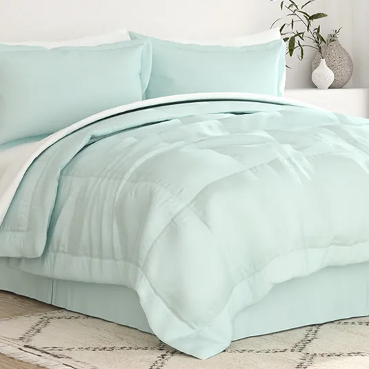 A light blue bedding set includes a comforter, two pillows, and a bed skirt, all displayed in a neatly arranged bedroom setting. A plant and vase are on a nearby surface, adding a decorative touch.
