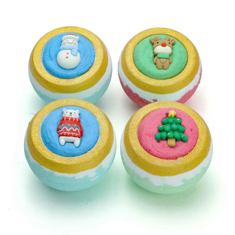 Four colorful holiday-themed bath bombs, each topped with a festive decoration: a snowman, a reindeer, a bear in a sweater, and a Christmas tree. They feature gold accents on their rims.