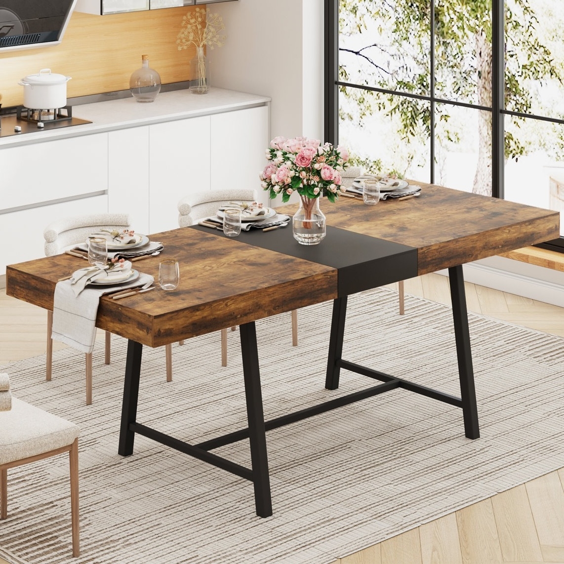 A modern dining table with a rustic wooden top and black metal legs is set for four, featuring plates, glasses, and napkins. A vase with pink flowers serves as the centerpiece. The setting is a bright kitchen with large windows and a neutral rug.