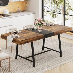 A modern dining table with a rustic wooden top and black metal legs is set for four, featuring plates, glasses, and napkins. A vase with pink flowers serves as the centerpiece. The setting is a bright kitchen with large windows and a neutral rug.
