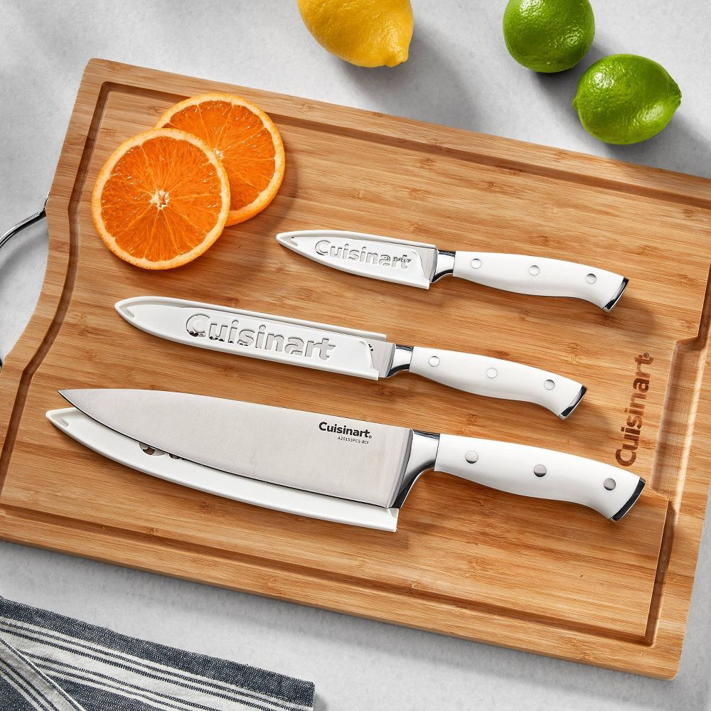 Three Cuisinart knives with white handles are displayed on a wooden cutting board, alongside sliced oranges, a lemon, and a lime.