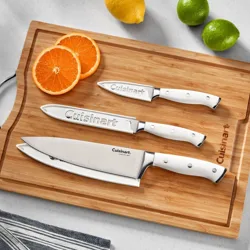 Three Cuisinart knives with white handles are displayed on a wooden cutting board, alongside sliced oranges, a lemon, and a lime.