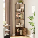 A corner bookshelf with four wooden shelves and black metal supports displays decorative items including flowers, framed art, books, and small sculptures. A potted plant stands nearby, enhancing the cozy corner setup.
