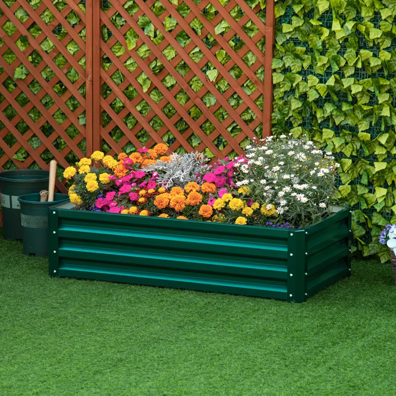 Green corrugated galvanized raised garden bed filled with colorful flowers, set against a wooden lattice and ivy backdrop. It includes marigolds, daisies, and other vibrant blooms, placed on a grassy area with gardening tools nearby.