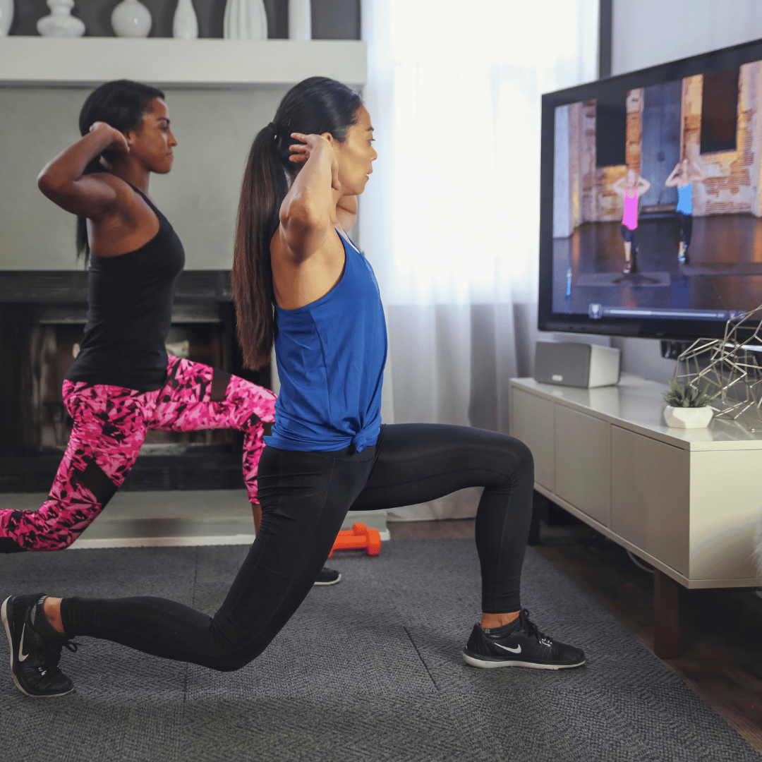 Two people are doing lunges while following a workout video on a wall-mounted TV. One is wearing a blue top and black leggings, the other a black top and pink patterned leggings. A small speaker and decorative items are on the white cabinet below the TV.