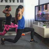 Two people are doing lunges while following a workout video on a wall-mounted TV. One is wearing a blue top and black leggings, the other a black top and pink patterned leggings. A small speaker and decorative items are on the white cabinet below the TV.