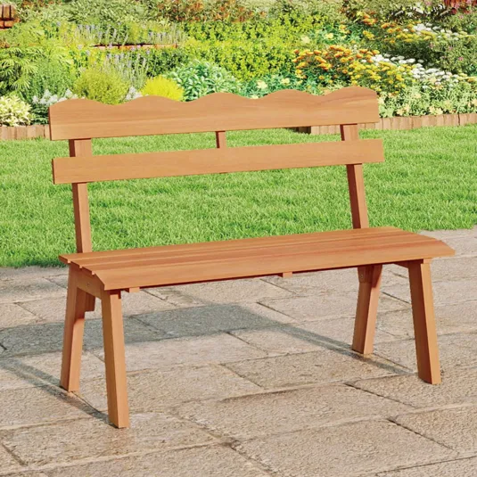 A wooden garden bench with a scalloped backrest is placed on a stone patio, surrounded by lush greenery and vibrant flowers.