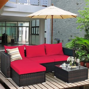 The 5-piece patio sectional set features dark brown wicker with vibrant red cushions, including a sofa and a matching coffee table with a glass top. It comes with an umbrella and a decorative striped pillow, set on a wooden deck surrounded by greenery.