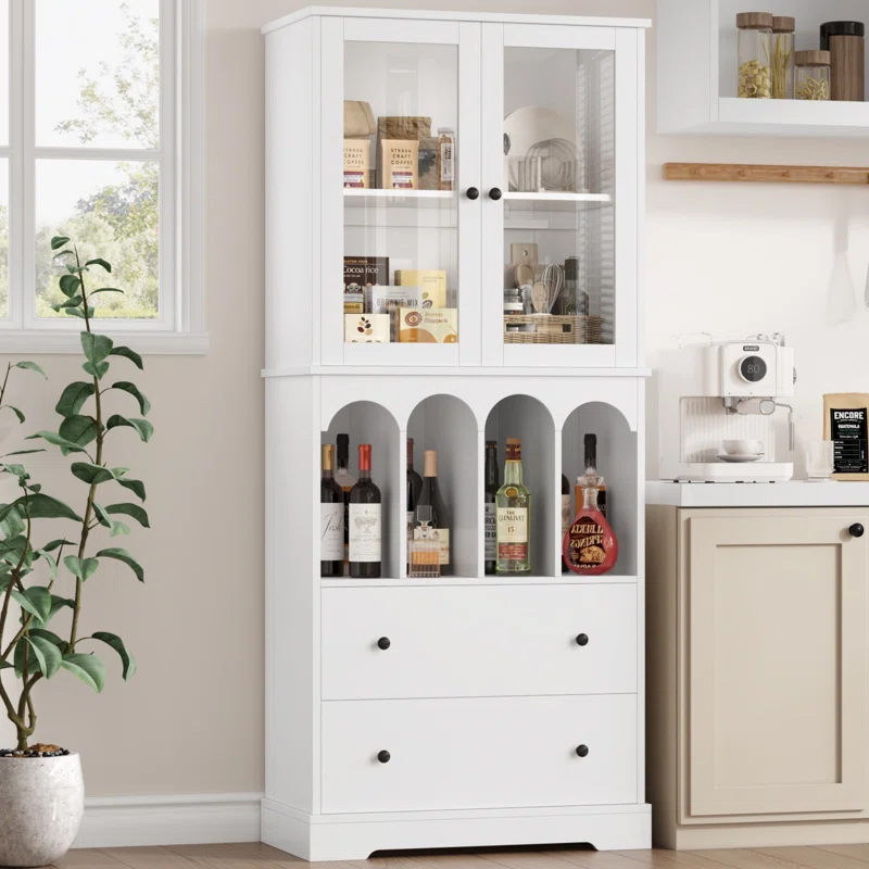 A white 71\“ kitchen pantry with a top section of glass doors displaying food items, a middle section with arches storing various bottles, and two bottom drawers. It's next to a coffee machine and accompanied by a potted plant.