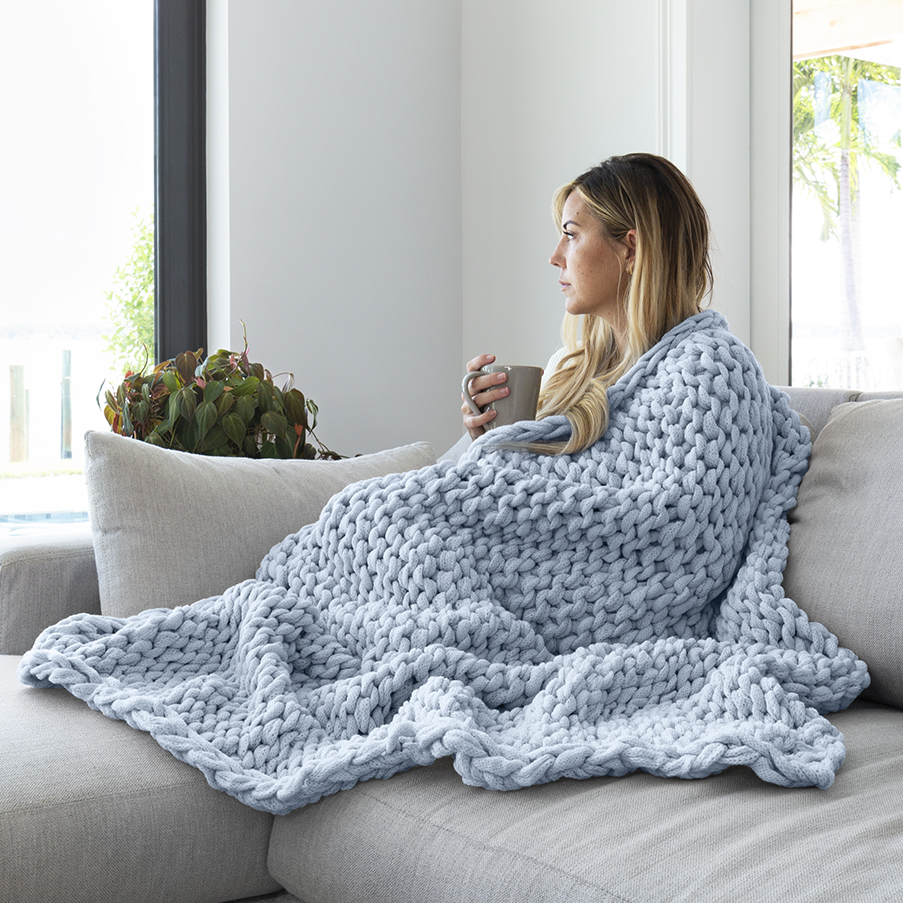 A woman is seated on a sofa, draped in a chunky knit blue blanket, holding a mug.