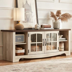 A rustic wooden sideboard features two glass-paneled cabinets and open shelves. It's decorated with a lamp, framed artwork, a small lantern, books, a decorative vase with dried pampas grass, and a small speaker.