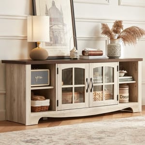 A rustic wooden sideboard features two glass-paneled cabinets and open shelves. It's decorated with a lamp, framed artwork, a small lantern, books, a decorative vase with dried pampas grass, and a small speaker.