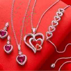 Silver jewelry set with heart-shaped designs includes two pendant necklaces, earrings, and a bracelet, all featuring red gemstones and diamond accents, displayed on a red backdrop.