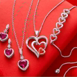 Silver jewelry set with heart-shaped designs includes two pendant necklaces, earrings, and a bracelet, all featuring red gemstones and diamond accents, displayed on a red backdrop.