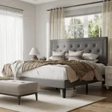 A bedroom features a gray tufted headboard bed with layered bedding in neutral tones, decorative pillows, a bedside table with a lamp, and a cushioned bench at the foot, all set in a well-lit room with large windows and sheer curtains.